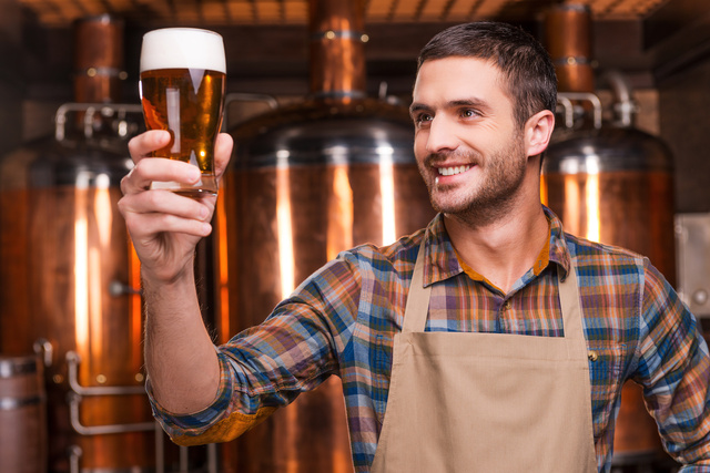 Happy brewer. Happy young male brewer in apron holding glass with beer and looking at it with smile while standing in front of metal containers
